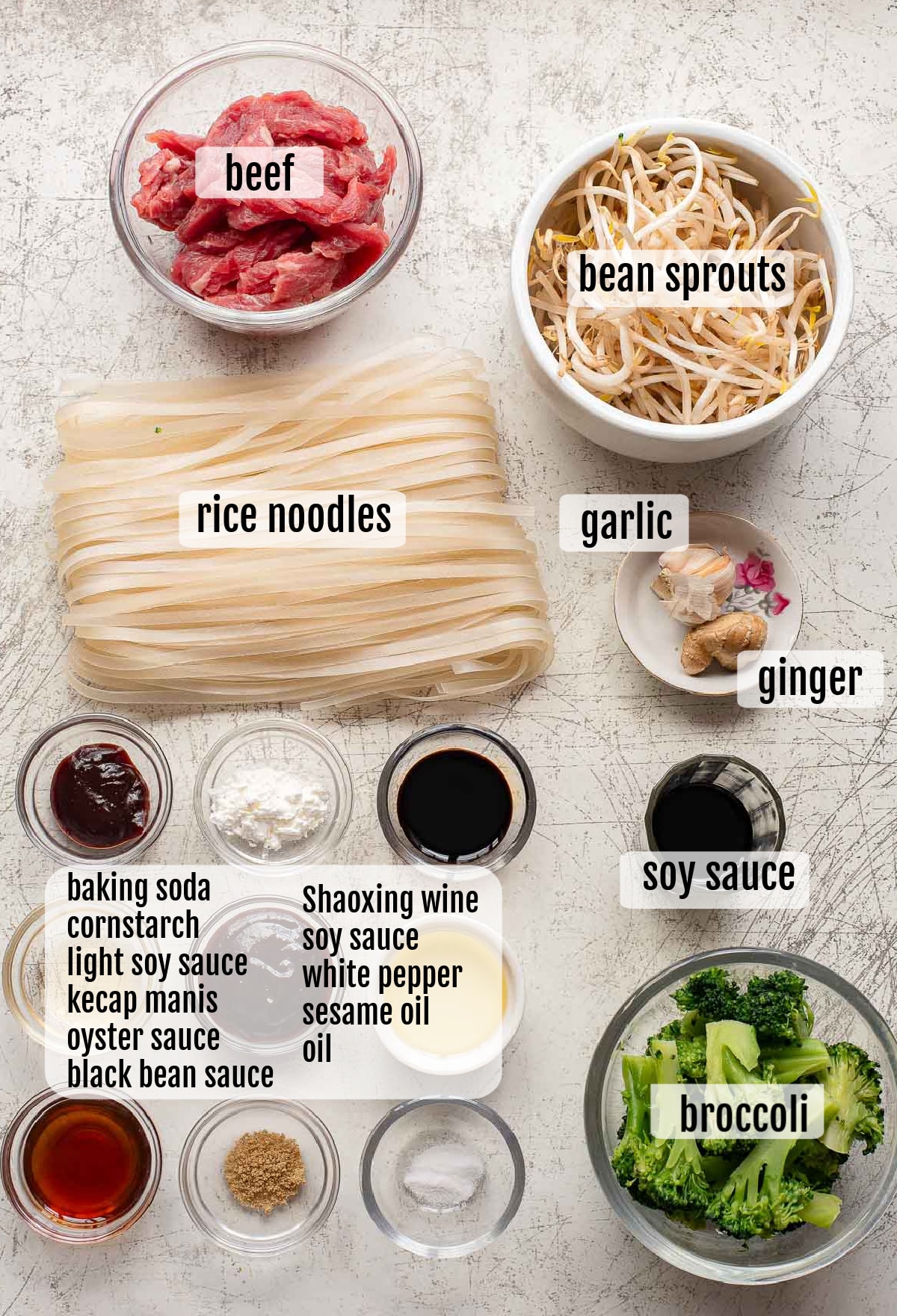 A top-down view of beef chow fun ingredients: sliced beef, bean sprouts, rice noodles, garlic, ginger, broccoli, various sauces, baking soda, cornstarch, and seasonings in bowls—each clearly labeled for easy stir-fry preparation.