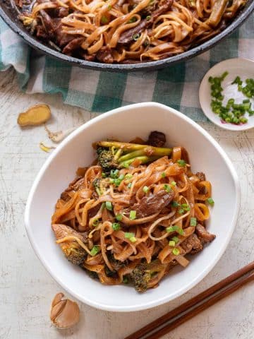 A bowl of beef chow fun—stir-fried rice noodles with beef, broccoli, and green onions—sits on a table beside chopsticks, garlic, and a small dish of sliced green onions. In the background, a skillet brims with more savory noodles.