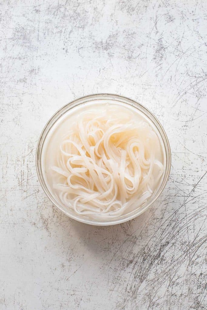 A glass bowl filled with soaked white rice noodles—perfect for making classic beef chow fun—sits on a lightly scratched, white textured surface.