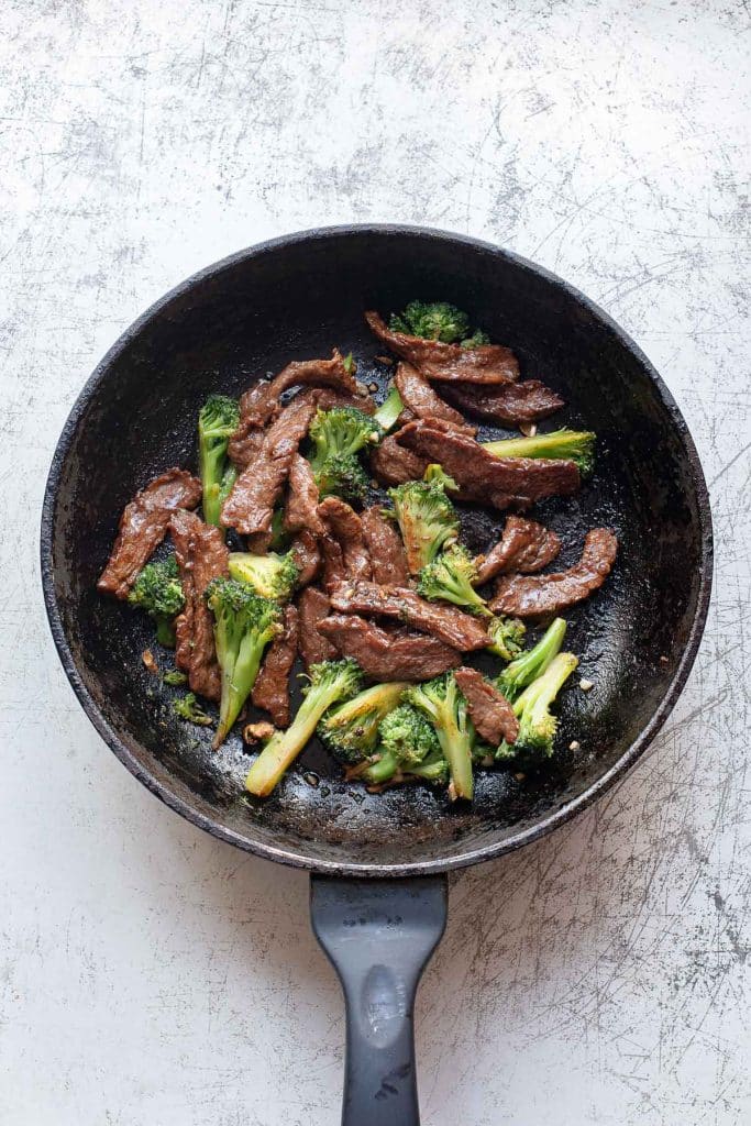 A black frying pan containing cooked beef strips and broccoli florets, reminiscent of beef chow fun, set on a scratched white surface. The beef appears browned and saucy, mixed with bright green broccoli.