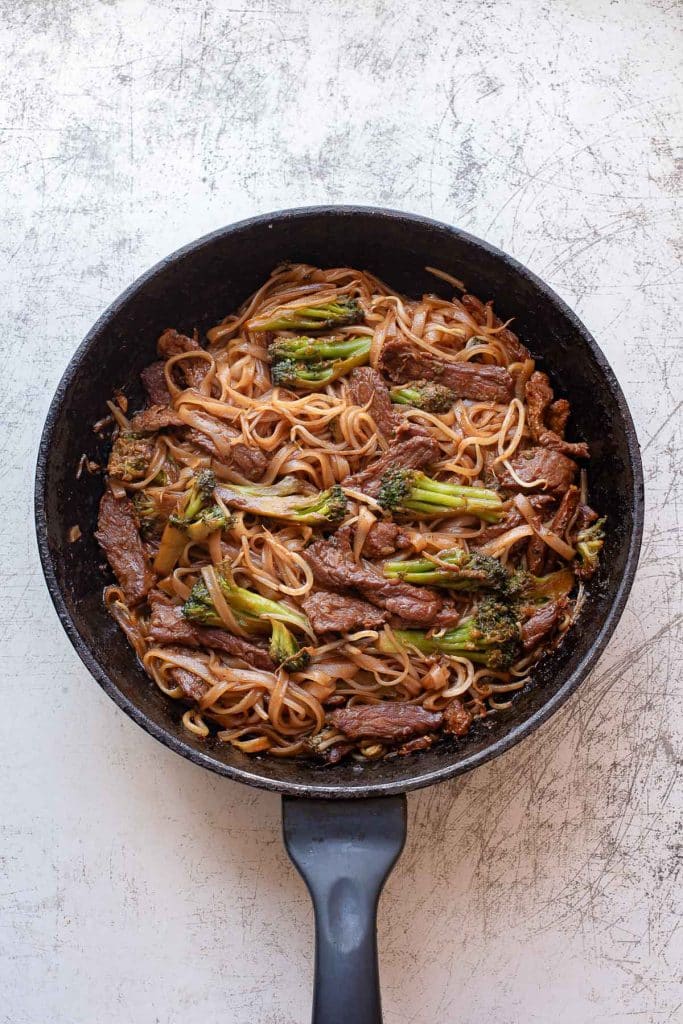 A black skillet filled with beef chow fun—stir-fried rice noodles, sliced beef, broccoli florets, and a dark savory sauce—rests on a light, textured surface.