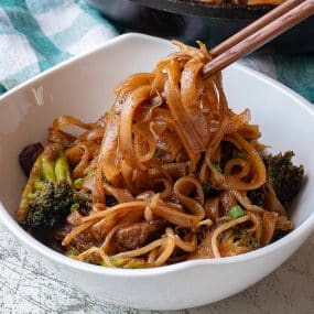A bowl of beef chow fun with stir-fried noodles, broccoli, and beef is lifted by wooden chopsticks. The noodles are coated in a dark sauce and served in a white bowl.