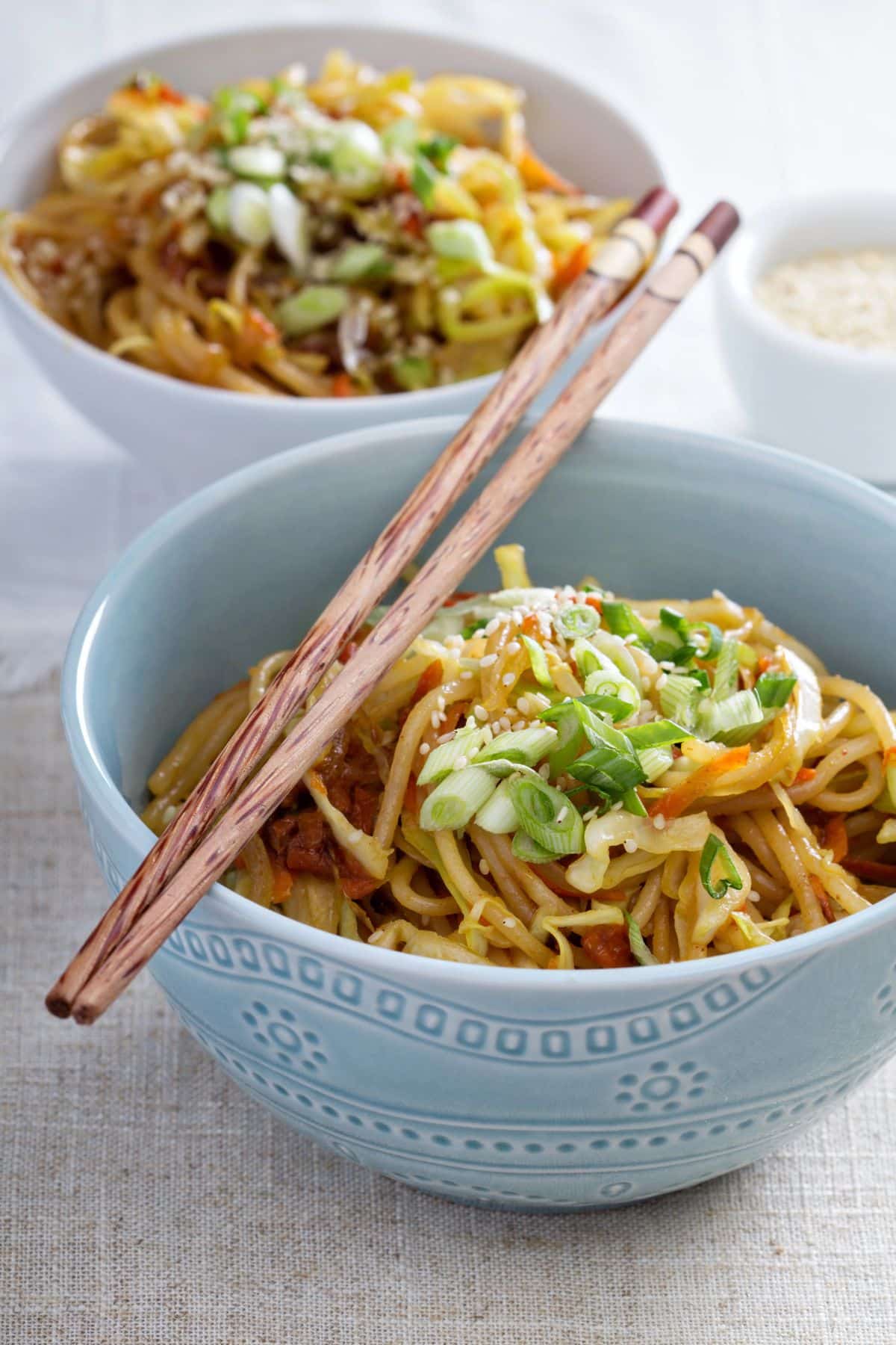 A close-up of two bowls of stir-fried noodles topped with chopped green onions and sesame seeds. One bowl is in the foreground, holding a pair of chopsticks resting on its rim, while the second bowl is in the background slightly out of focus, with a small dish of sesame seeds nearby.
