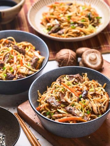 A table set with three bowls of stir-fried noodles mixed with vegetables, mushrooms, and pieces of meat. A pair of chopsticks and a small dish of sesame seeds are placed beside the bowls. Behind the main dishes, there is a ceramic bowl containing a sauce.