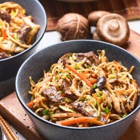 A bowl of stir-fried noodles mixed with sliced mushrooms, carrot strips, and green onions. Another bowl with a similar dish is partially visible in the background. Two whole mushrooms and some chopsticks are placed beside the bowls on a wooden surface.