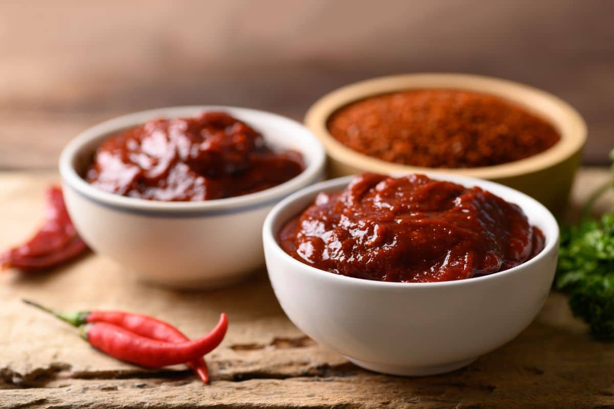 Two white bowls filled with rich, dark red chili paste are placed on a wooden surface. In the background, a wooden bowl of red chili powder can be seen. Beside the bowls, there are whole red chili peppers and some green herbs.
