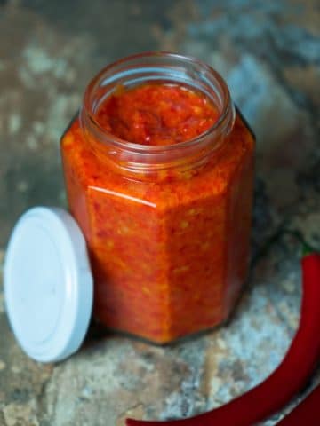 A hexagonal glass jar filled with vibrant red chili paste is placed on a rustic surface. The jar's white lid is off and lies beside it. Two whole red chili peppers are also laid next to the jar, emphasizing the spicy nature of the contents.