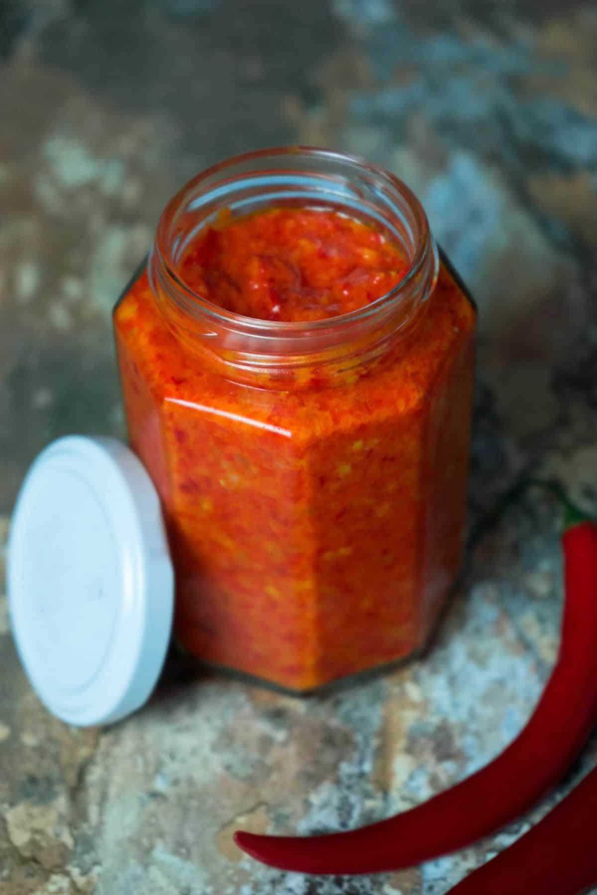 A hexagonal glass jar filled with vibrant red chili paste is placed on a rustic surface. The jar's white lid is off and lies beside it. Two whole red chili peppers are also laid next to the jar, emphasizing the spicy nature of the contents.