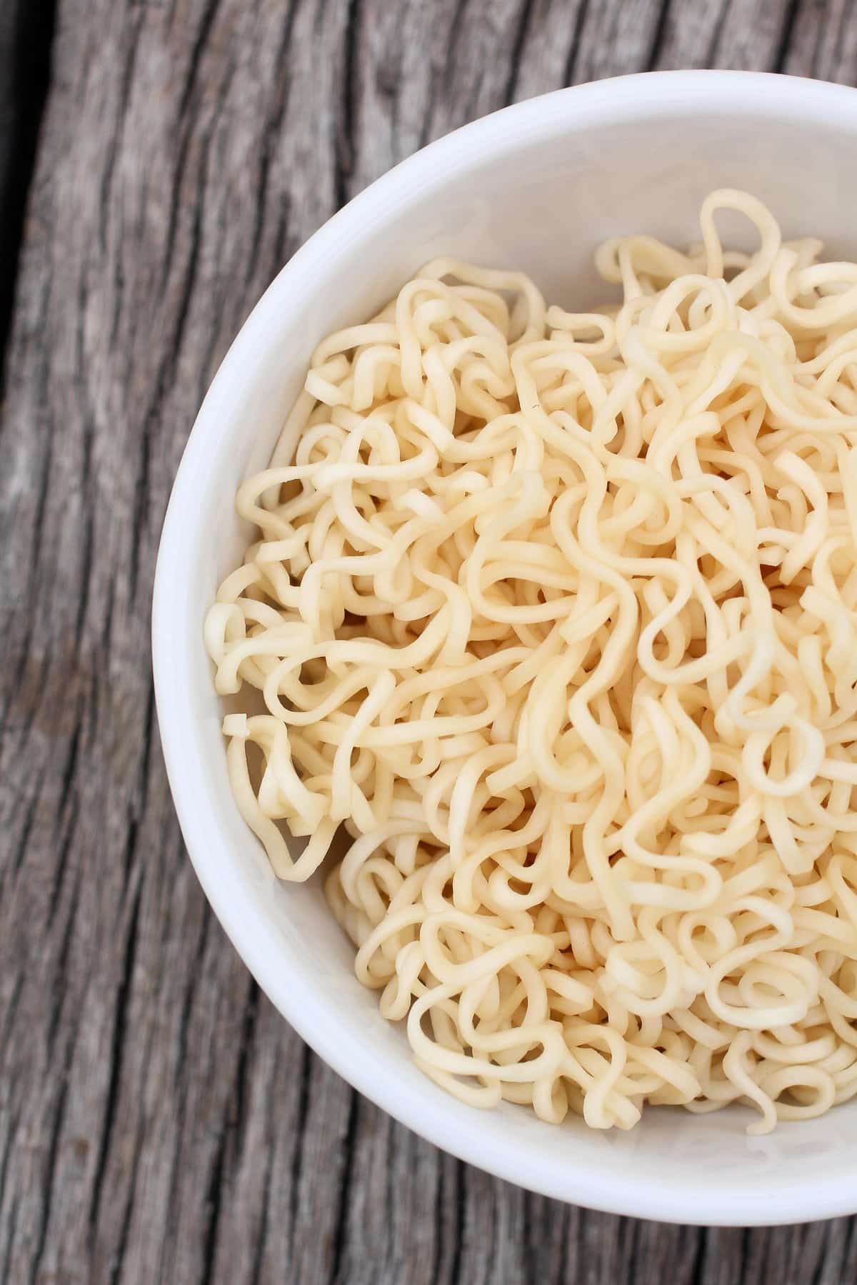A close-up shot of a white bowl filled with curly, dry Asian noodles, placed on a textured wooden surface. The instant ramen noodles appear uncooked and neatly arranged within the bowl.
