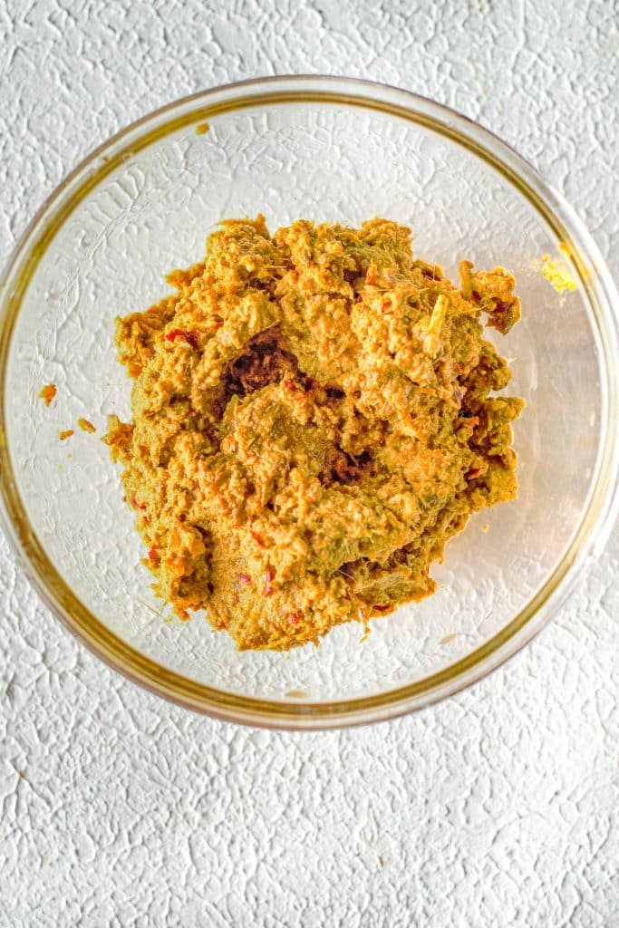 A clear glass bowl containing a mixture of various ground spices and ingredients, forming a brownish paste, is placed on a textured white surface.
