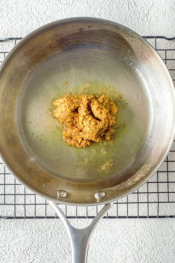 A stainless steel pan sits on a wire cooling rack with a small heap of browned, grainy seasoning or ground spice blend in the center. The background is a textured, light-colored surface.