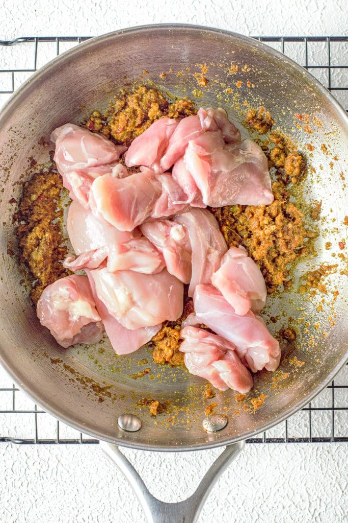 A metal skillet on a wire rack contains chopped raw chicken pieces on top of a mixture of minced seasonings and spices. The skillet has a well-worn appearance, and the background surface is textured.
