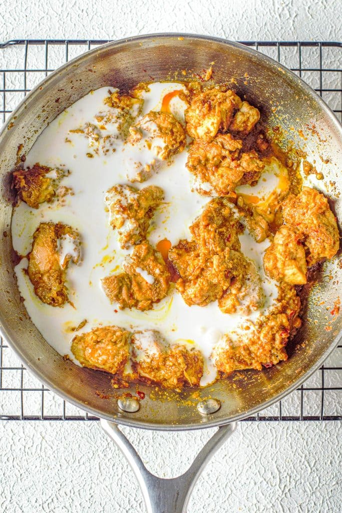 A stainless steel skillet on a cooling rack contains pieces of cooked chicken covered in a rich, thick, golden-brown sauce with white creamy coconut milk drizzled on top. The skillet sits on a textured white surface.