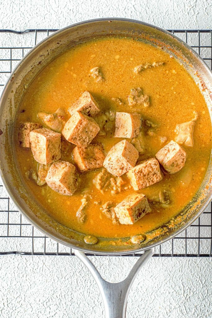 A skillet is filled with a rich orange curry sauce and chunky cubes of tofu. The sauce appears to be seasoned and thick, surrounding the tofu pieces, which are lightly browned. The skillet is placed on a wire rack over a textured white surface.