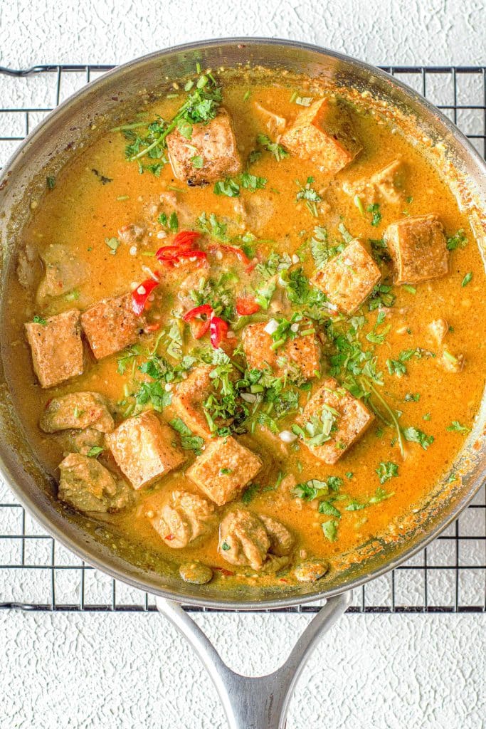 A round skillet filled with a vibrant curry dish, featuring rectangular tofu pieces, slices of red chili, chopped cilantro, and a golden-orange sauce. The skillet is on a cooling rack against a textured white surface.