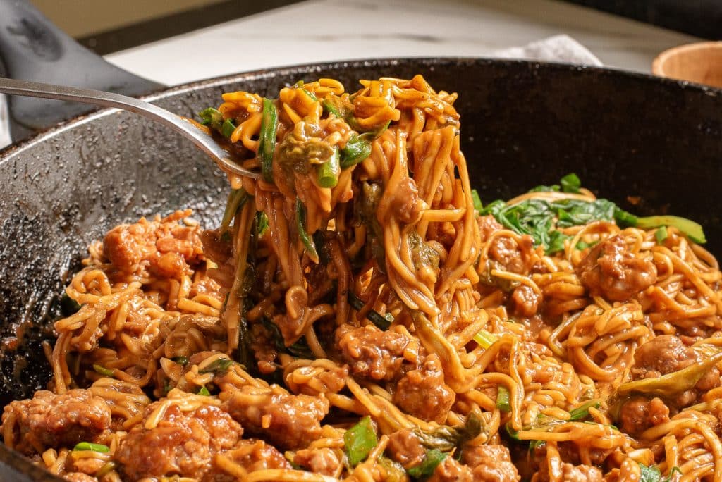 A skillet filled with cooked noodles, green vegetables, and pieces of seasoned meat. A spoon is lifting a portion, showcasing the savory sauce coating the ingredients. The dish appears to be a hearty, savory stir-fry.