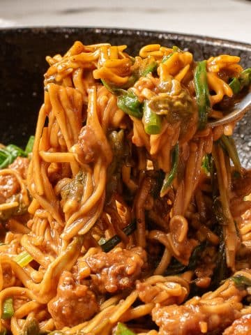 A fork lifts a tantalizing forkful of noodles mixed with sausage and vegetables from a black skillet. The dish appears to be creamy and richly seasoned, reminiscent of dan dan noodles, with visible bits of meat and greens intertwined with the noodles.
