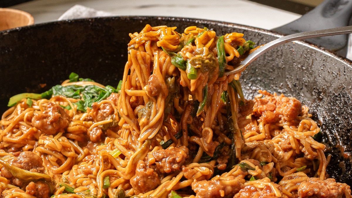 A fork lifts a tantalizing forkful of noodles mixed with sausage and vegetables from a black skillet. The dish appears to be creamy and richly seasoned, reminiscent of dan dan noodles, with visible bits of meat and greens intertwined with the noodles.
