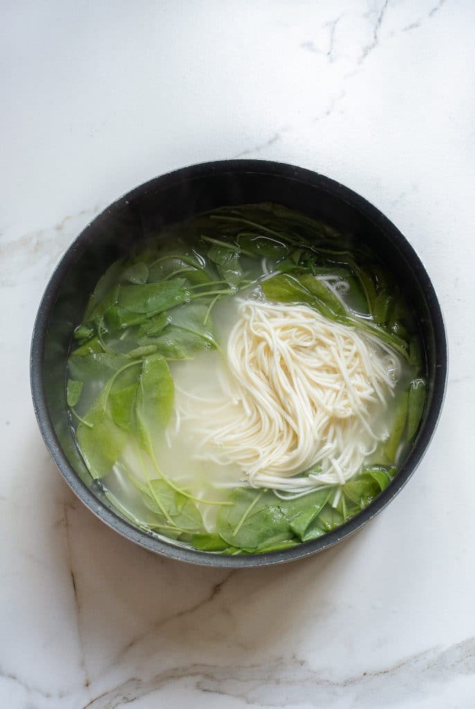 A black pot on a white marble surface contains a steaming broth with leafy greens and a bundle of thin dan dan noodles. The leafy greens float around the partially submerged noodles, adding a touch of color to the hot, aromatic dish.