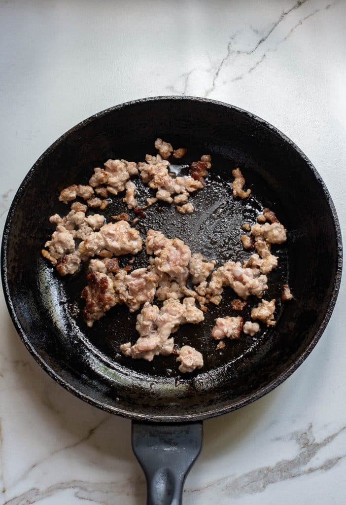A black frying pan containing partially cooked ground meat sits on a white marble countertop. The meat, reminiscent of a key ingredient in dan dan noodles, is slightly pink with some browned bits, indicating it's in the middle of the cooking process. The pan shows signs of use with some oil residue.