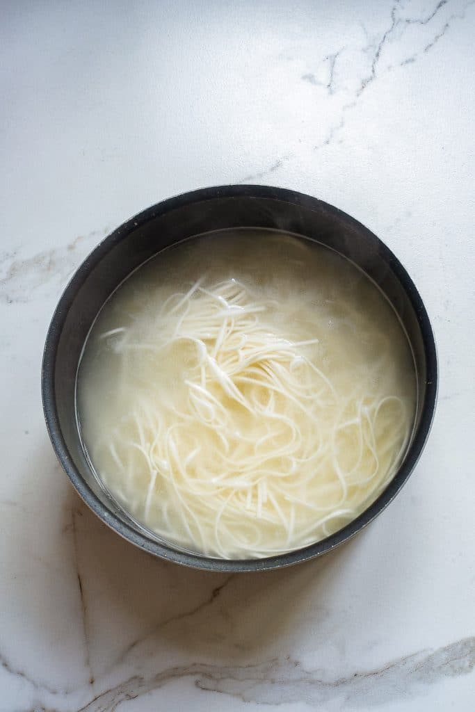 A black bowl is filled with steaming hot broth and thin white dan dan noodles. The bowl is placed on a light-colored marble surface. The broth appears milky, and the noodles are slightly tangled. The image conveys a warm and comforting meal.
