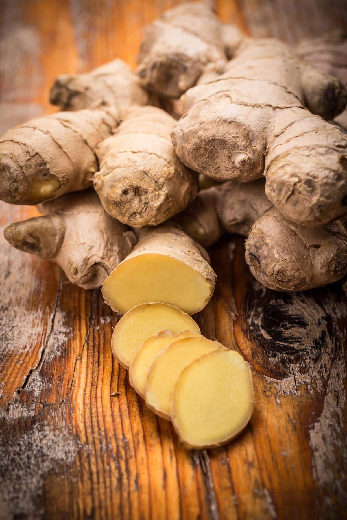 Fresh ginger roots stacked on a rustic wooden surface with a few slices of ginger in the foreground, displaying the yellowish interior contrasted against the rough, brown skin of the root.