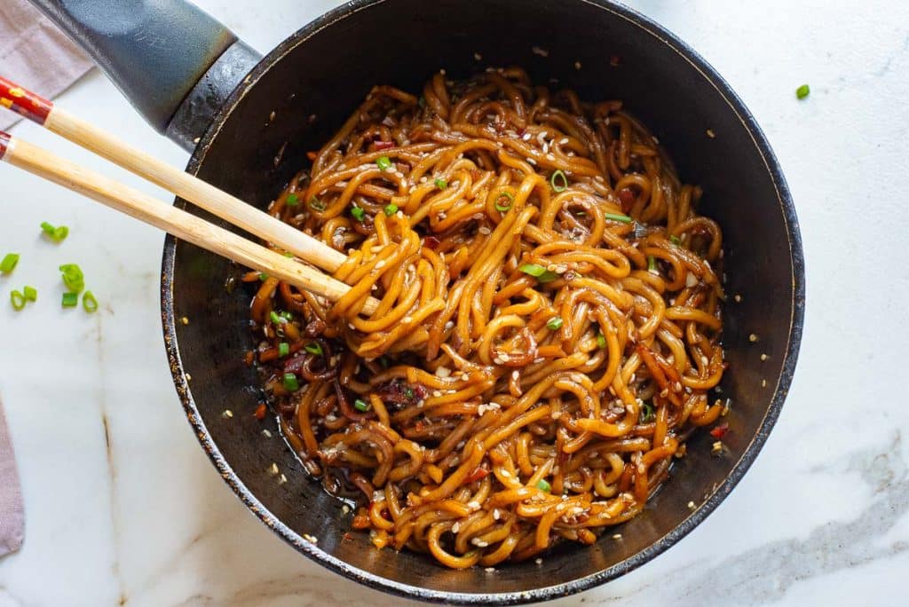 A black pan filled with garlic chili oil noodles, garnished with chopped green onions and sesame seeds. Two wooden chopsticks are picking up a portion of the noodles. The background features a white marble surface with a hint of a pink napkin on the side.