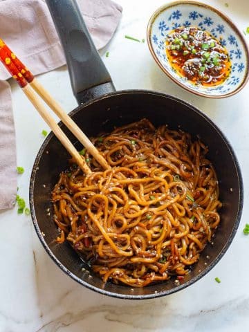 A pan filled with garlic chili oil noodles mixed in a rich, dark sauce is presented on a white marble surface. Wooden chopsticks rest on the edge of the pan. A small blue and white dish containing spicy chili sauce with green garnishes is placed beside the pan.