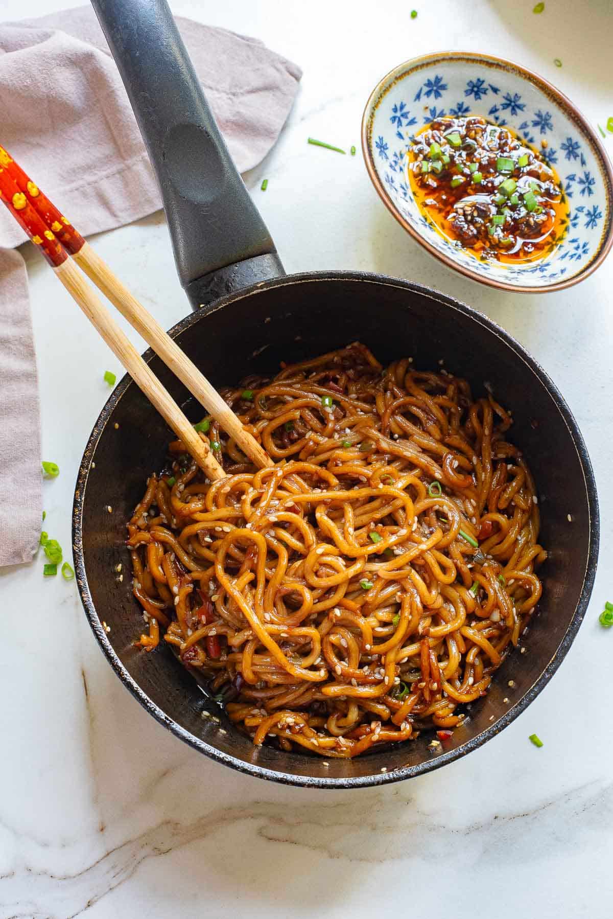 A pan filled with garlic chili oil noodles mixed in a rich, dark sauce is presented on a white marble surface. Wooden chopsticks rest on the edge of the pan. A small blue and white dish containing spicy chili sauce with green garnishes is placed beside the pan.