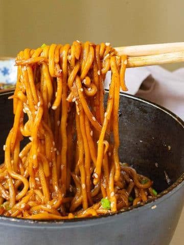 A person holds chopsticks lifting a portion of garlic chili oil noodles from a black wok. The noodles are coated in sauce and mixed with vegetables, and the background shows a white surface with a partially visible plate and napkin.
