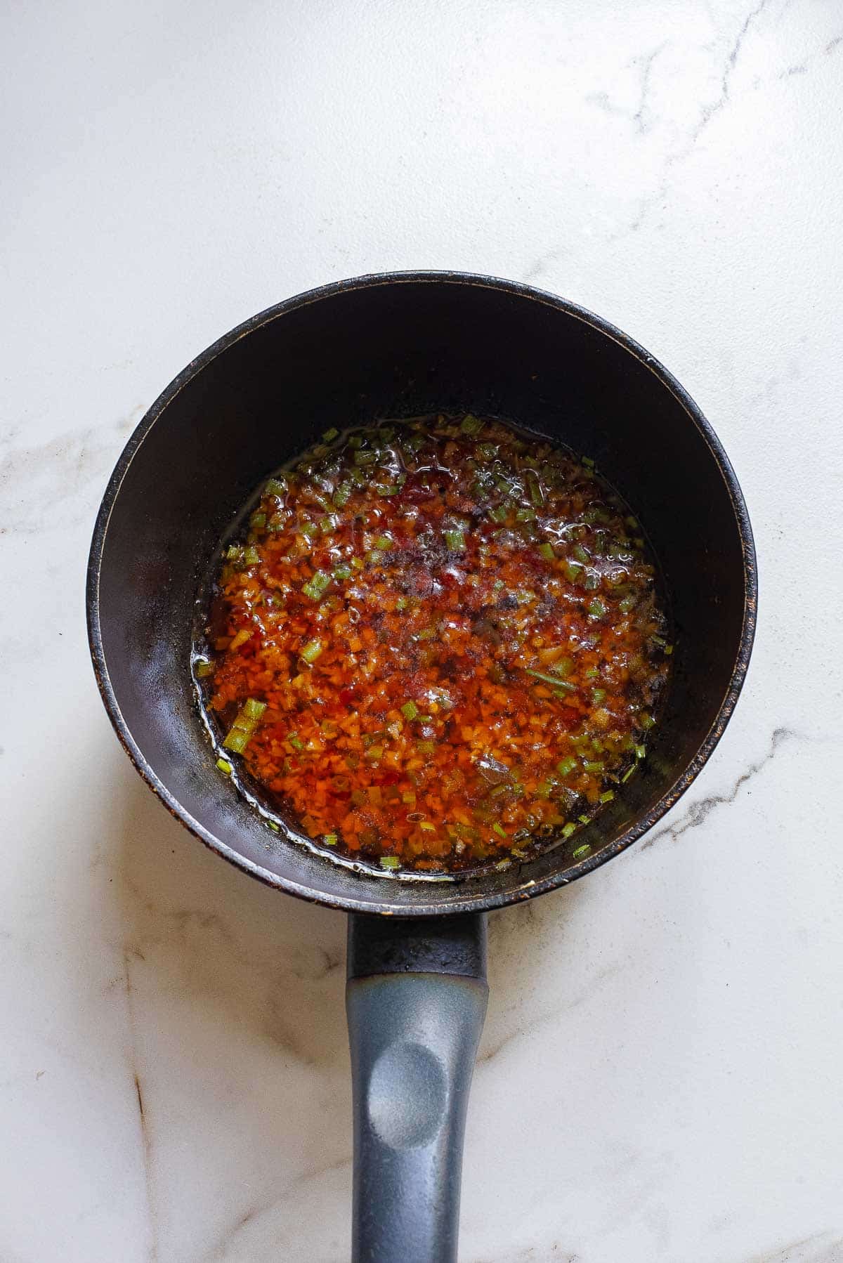 A black saucepan containing a simmering mixture of minced vegetables and herbs in a vibrant red sauce with hints of garlic chili oil sits on a white marble countertop. The sauce appears to be evenly mixed with small pieces of green, orange, and yellow vegetables.