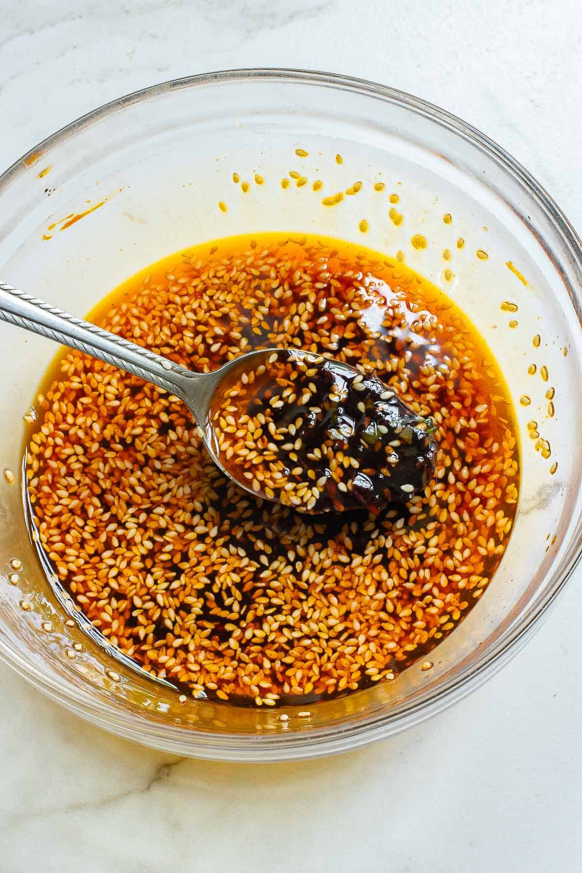 A spoon held over a transparent bowl filled with sesame seeds and dark soy sauce, along with a light-colored liquid, evokes the rich aroma of garlic chili oil. The deep brown sauce is speckled with sesame seeds throughout, set against a pristine white countertop.