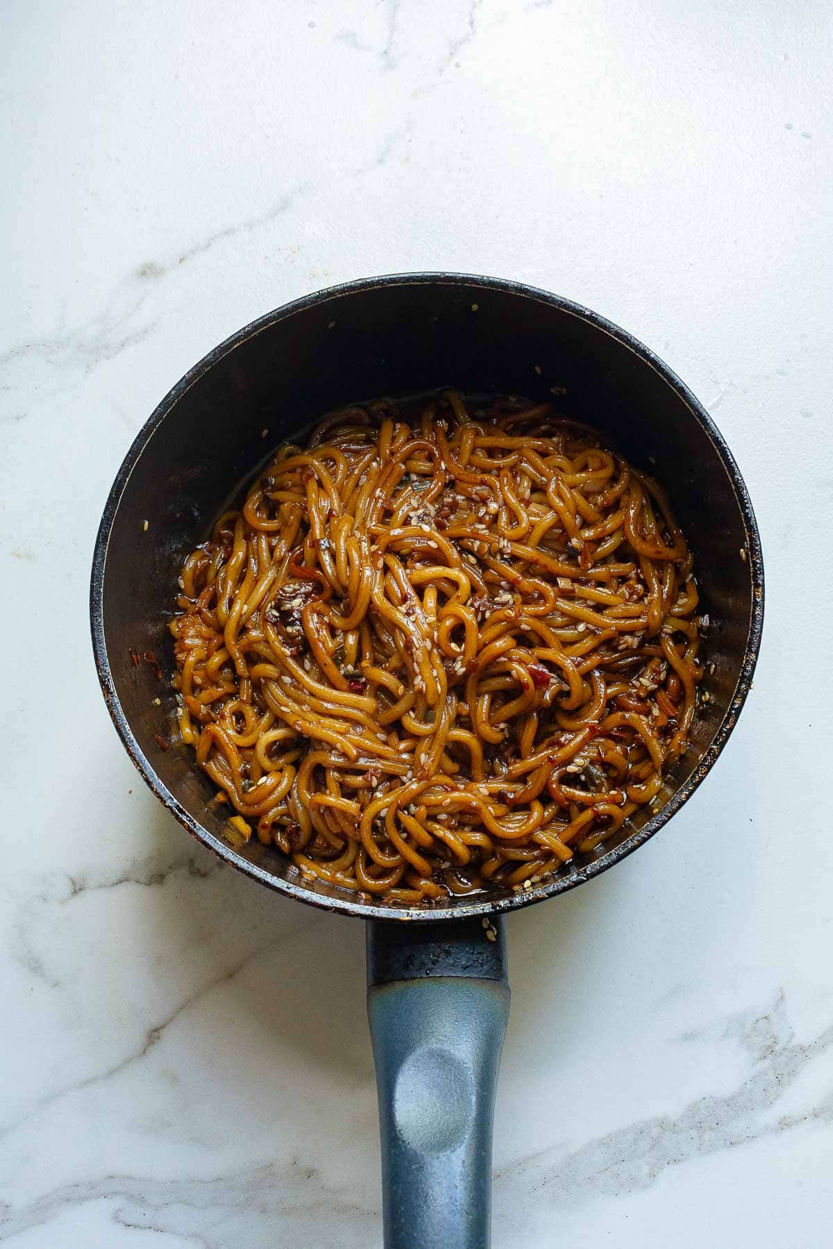 A pot filled with cooked noodles mixed in a dark savory sauce, enhanced by garlic chili oil, with visible bits of vegetables and spices, sitting on a white marble countertop.