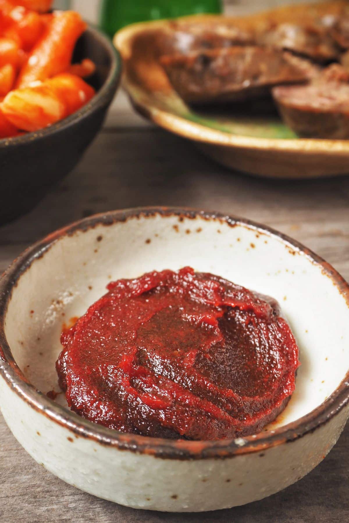 A ceramic bowl filled with a dollop of red chili paste is in focus. The bowl is placed on a wooden surface. In the background, another bowl contains orange pickled vegetables, and a plate has meat slices. For those wondering what is gochujang, it might be that tantalizing red paste in the foreground.