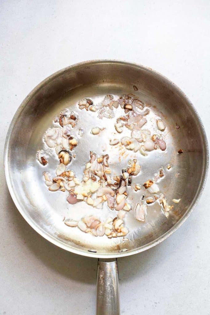A stainless steel frying pan with bits of sautéed garlic and onion, some of which are slightly browned, sits on a light-colored surface.