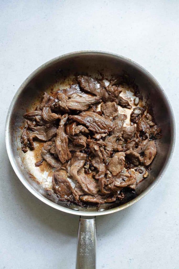 A stainless steel skillet containing cooked, sliced beef strips on a light gray surface. The meat appears to be well-browned and seasoned, with some visible residue from cooking.
