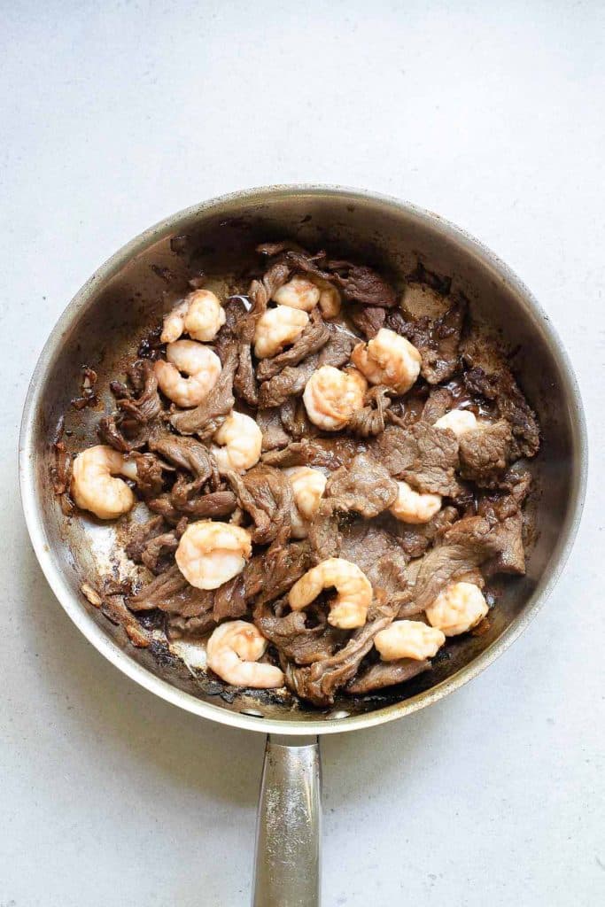 A frying pan contains cooked shrimp and pieces of beef. The shrimp are pink and curled, while the beef is thinly sliced and browned. The surface beneath the pan is a light gray countertop.