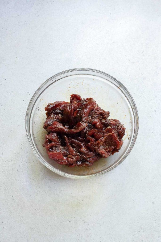A clear glass bowl filled with marinated strips of raw beef sits on a light gray surface. The beef appears to be seasoned with dark spices. The ingredients are well-mixed and ready for cooking.