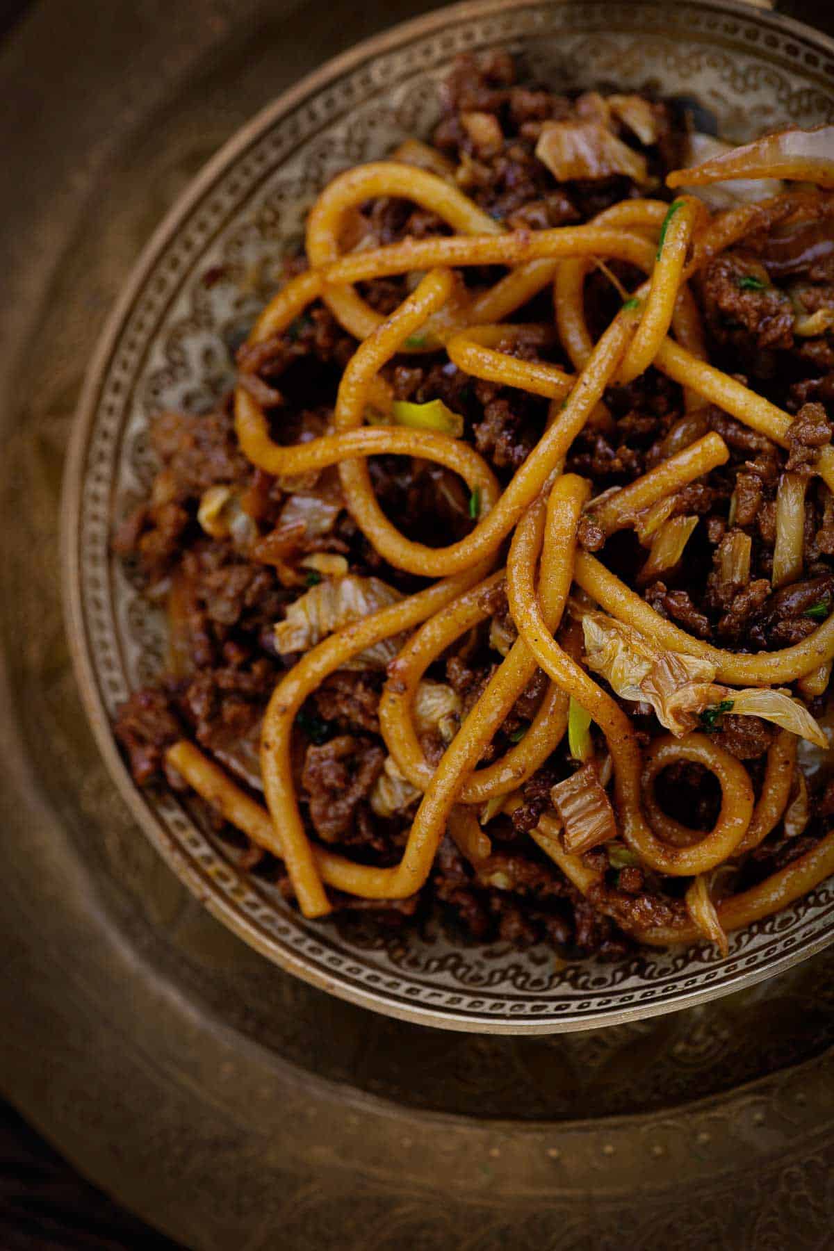 A close-up of a bowl of stir-fried noodles mixed with ground meat, shredded cabbage, and green onions. The dish has a rich, dark sauce, and the noodles are thick and slightly glossy. The bowl is ornate, with intricate patterns on the rim.