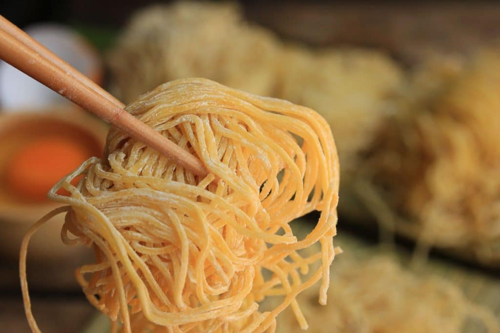 Close-up of chopsticks holding a bundle of uncooked yellow noodles. The background is slightly blurred, showing more piles of similar noodles and a bowl with an orange ingredient, possibly an egg yolk. The texture of the noodles appears dry and slightly floury.
