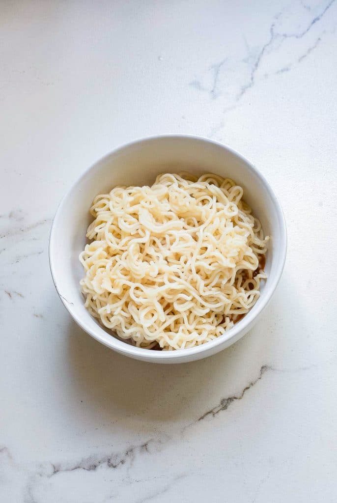 A white bowl filled with plain cooked noodles is placed on a white marble countertop. The noodles are tightly packed and have a soft, slightly curly appearance.