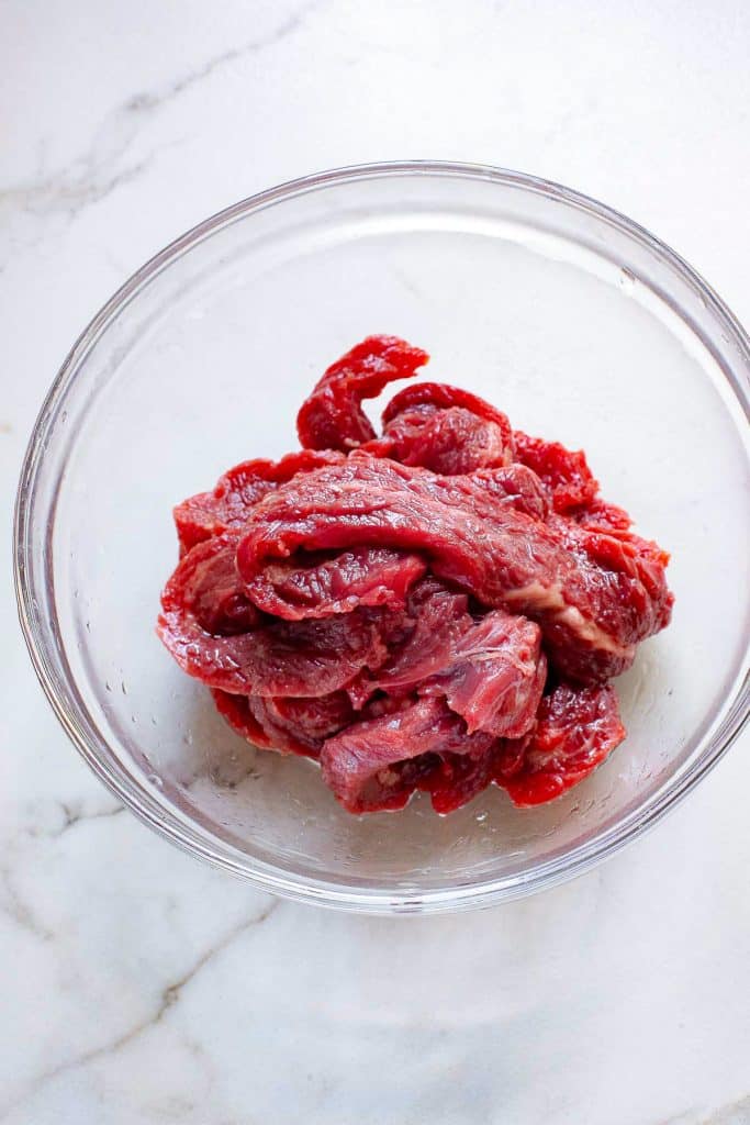 A clear glass bowl filled with raw, red beef slices sits on a white marble countertop. The meat appears fresh and ready for cooking.