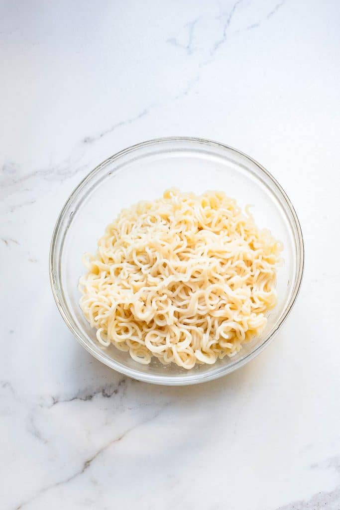 A clear glass bowl filled with cooked instant ramen noodles is placed on a white marble surface. The noodles are lightly twisted and appear soft and ready to eat.