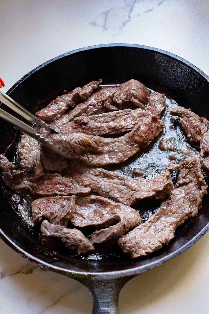 A cast iron skillet on a stove contains several strips of cooked beef. A pair of tongs, held by an unseen person, is grasping one of the beef strips. The skillet and beef are on a white countertop.