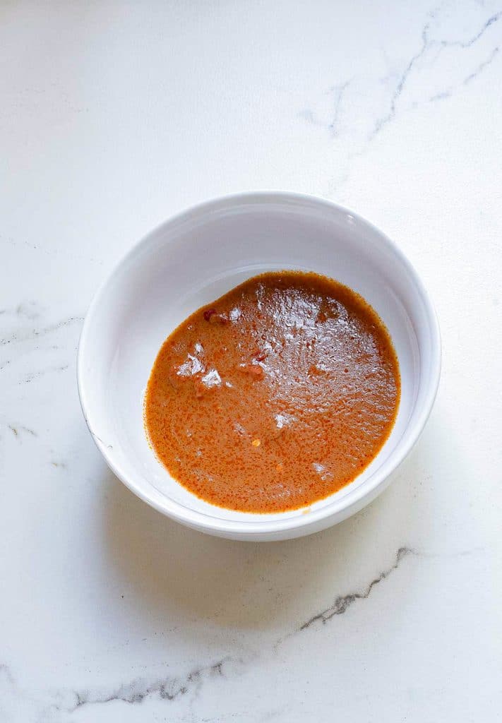 A white bowl containing a partially smooth, partially chunky reddish-brown sauce sits on a white marble surface. The sauce appears to have a blend of spices and a thin consistency.
