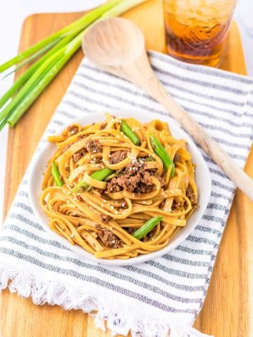 A plate of beef lo mein garnished with green onions and sesame seeds sits on a white and gray striped cloth atop a wooden board. Next to the plate is a wooden spoon and a glass of iced tea. Green onions are also placed on the board.