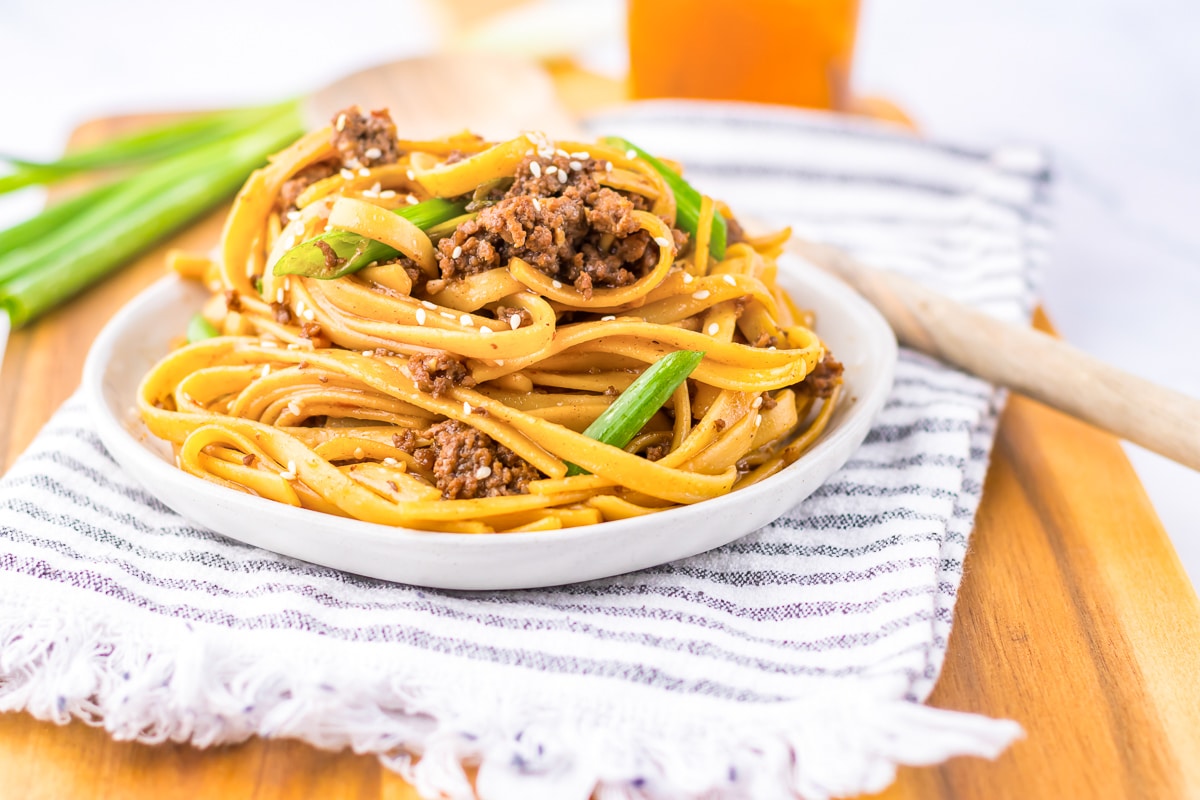 A plate of Asian-style noodles topped with ground meat, sliced green onions, and sesame seeds sits on a striped cloth napkin on a wooden cutting board. A pair of chopsticks rests beside the plate, and a glass of iced tea is in the background.
