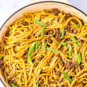 A close-up of a pan filled with cooked fettuccine pasta mixed with ground meat, topped with sesame seeds and garnished with chopped green onions. The pasta is coated with a shiny sauce that adds a rich color to the dish.