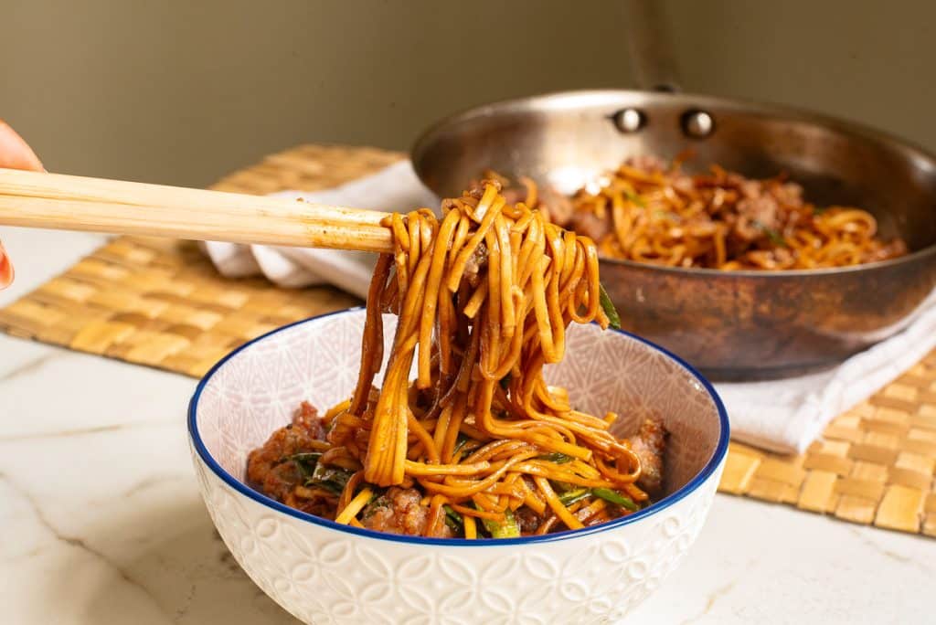 A close-up of chopsticks lifting stir-fried noodles with vegetables and meat from a white bowl with an intricate pattern. A pan with the same noodle dish and a placemat are in the background. The dish is saucy and garnished with greens.