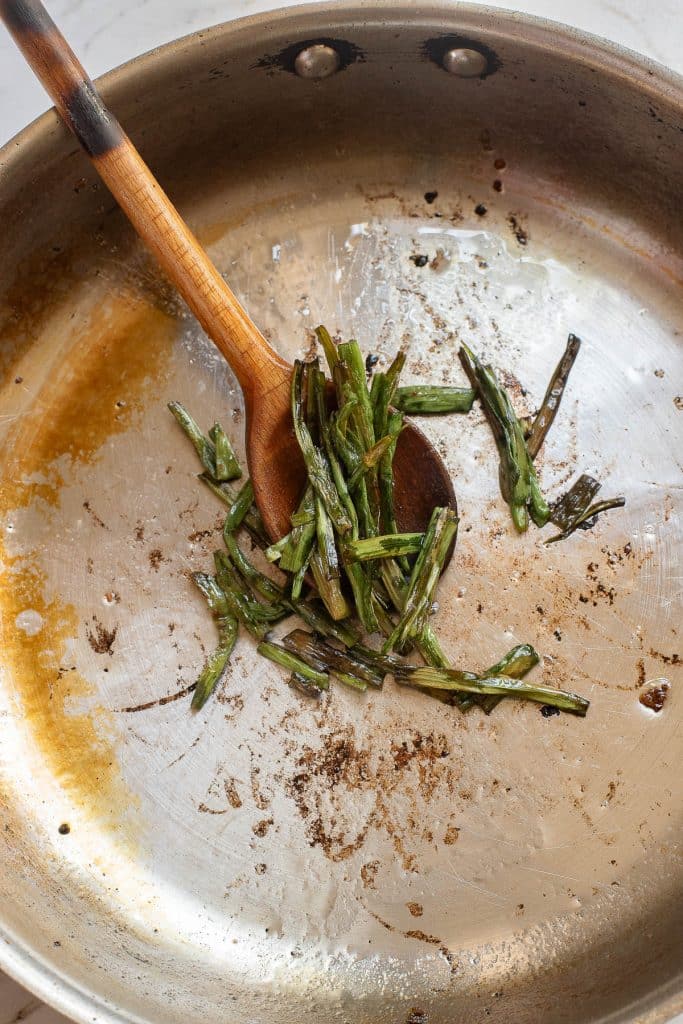 A metal frying pan contains partially cooked green stalks, likely a vegetable, with a minimal amount of oil and browned residue. A wooden spoon rests on the pan, slightly lifting some of the stalks. The pan shows signs of use with scattered browned bits.